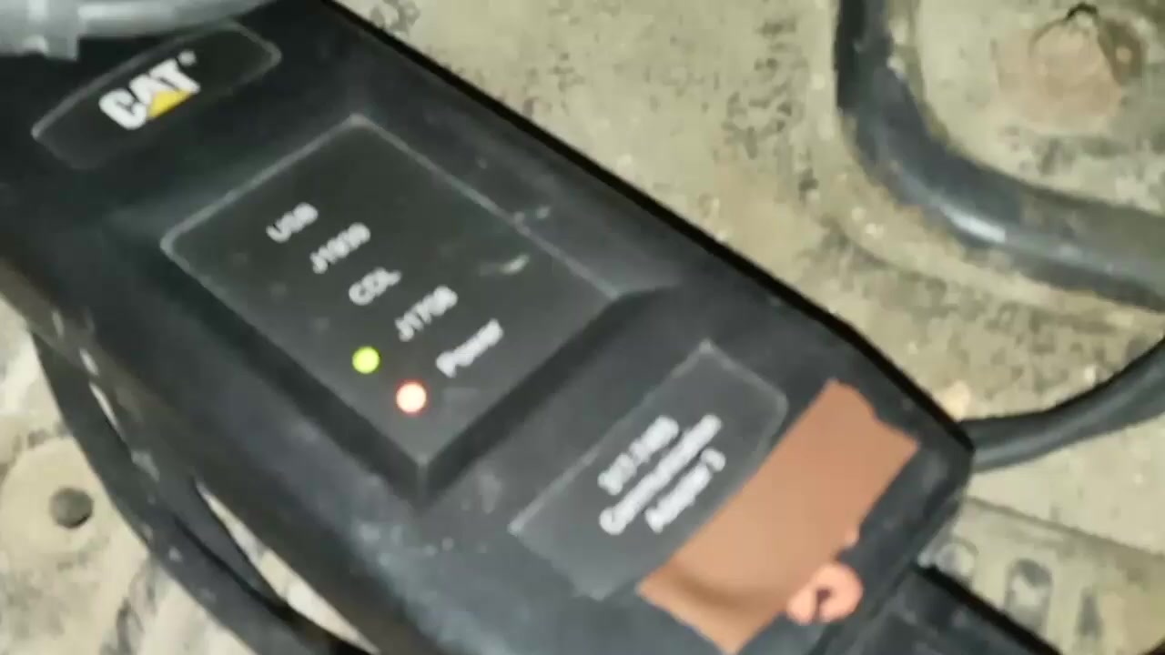 Close-up of a CAT Communication Adapter 3 connected to a vehicle's diagnostic port, with power and communication lights illuminated.